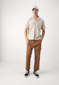 Camicia beige a maniche corte con bottoni e tasche sul petto, abbinata a pantaloni marroni e sneakers nere con suole e lacci bianchi.