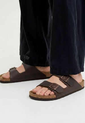 Voeten met bruine leren dubbele riem sandalen met gespen en kurken zolen, gecombineerd met losse zwarte broeken.