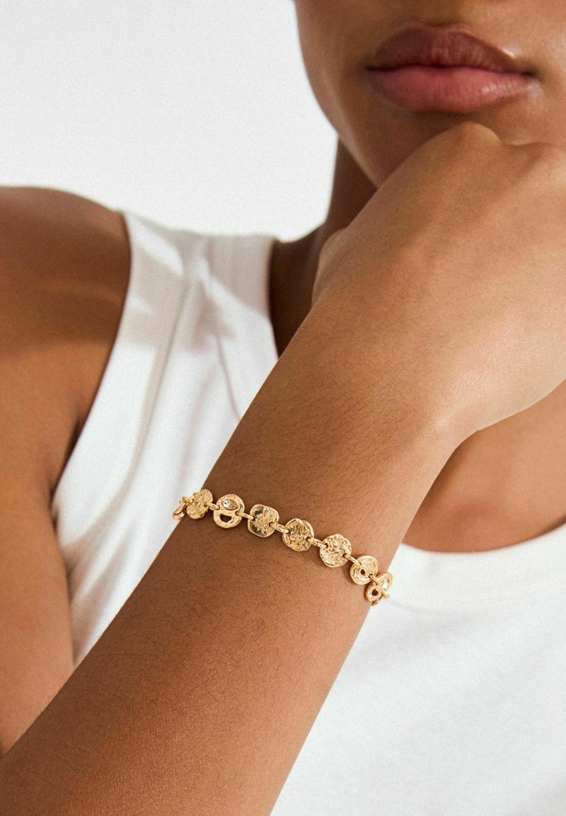 Bracelet en or composé de maillons circulaires à motifs texturés. Porté sur un poignet nu, associé à un haut blanc sans manches.