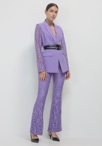 Completo due pezzi su misura di un delicato viola. La giacca presenta maniche in pizzo; i pantaloni aderenti mostrano anch'essi motivi in pizzo. Una cintura nera accentua la vita.