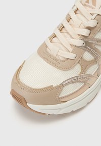 Zapatilla de color beige claro con detalles de tela de malla y cuero. Presenta cordones planos, un cuello acolchado y una suela de goma texturizada.