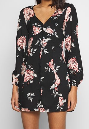 Vrouw draagt een zwarte jurk met lange mouwen, bedrukt met roze en witte bloemen, V-hals en knopen aan de voorkant.