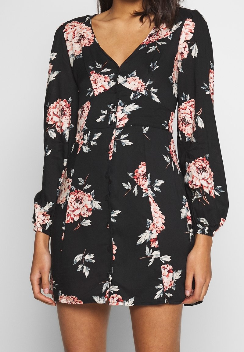 Femme portant une robe noire à manches longues avec des fleurs roses et blanches, un décolleté en V et des boutons sur le devant.