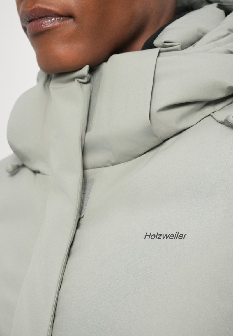 Veste isolante gris clair avec un col haut, présentant une texture lisse et un branding subtil "Holzweiler" sur la poitrine.