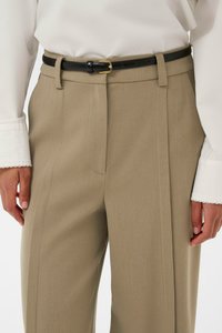 Bežīgas šūtas bikses ar teksturētu virsmu, kurām ir melna ādas josta ar zelta sprādzi, apvienotas ar baltu garām piedurknēm kreklu.