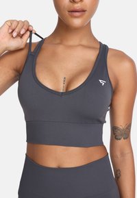 Czarny sportowy stanik w ciemnoszarym kolorze z głębokim dekoltem w kształcie litery V, szerokimi paskami i regulowanymi ramiączkami. Posiada akcent logo oraz gładki, elastyczny materiał.