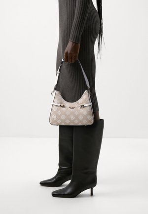 Beige handtas met patroon en een gebogen vorm, witte band en zilverkleurige beslag, vastgehouden door een persoon in een grijze ribgebreide jurk en zwarte laarzen tot aan de knie.