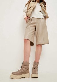 Shorts de cuero sintético beige con un corte relajado, que presentan pliegues frontales. Complementados con botas de tobillo acolchadas en tono marrón claro con suela gruesa y cremallera.