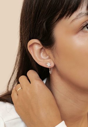 Silberne Blumenohrringe mit einer hängenden Kette und kleinen Edelsteinen, getragen am Ohr, eine Hand berührt sanft den Hals. Heller Hintergrund.