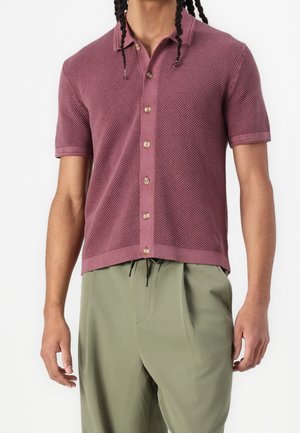 Hombre con camisa de manga corta texturizada color malva con botones y pantalones sueltos de color verde oliva claro sobre un fondo liso.