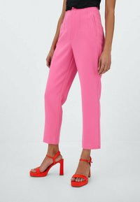 Pantalon rose sur mesure à taille haute avec poches latérales, associé à des talons blocs rouges ouverts à l'avant avec des brides à la cheville.