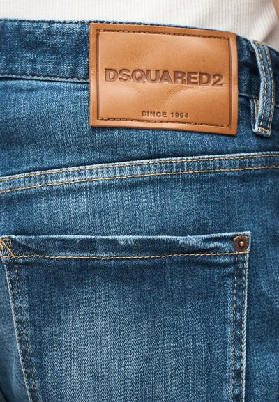 Poche arrière de jean en denim bleu avec étiquette de marque en cuir "DSQUARED2" et texte "Since 1964" cousu au-dessus de la poche.