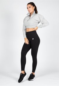 Grijze cropped sweatshirt met een halve ritssluiting en geribbelde boorden, gecombineerd met hoogslag zwarte leggings met een witte logo-detail. Zwarte sportschoenen.