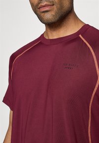 Κάτω αθλητικό μπλουζάκι σε μπορντό με πορτοκαλί ραφές. Διαθέτει στρογγυλή λαιμόκοψη και εκτύπωση λογότυπου στο στήθος, κατασκευασμένο από ελαφρύ ύφασμα.