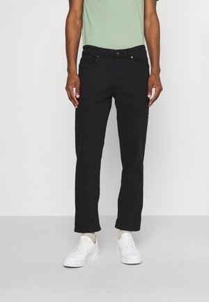 Mężczyzna ubrany w czarne jeansy o prostych nogawkach, jasnozieloną koszulkę i białe trampki, stojący na jednolitym tle.