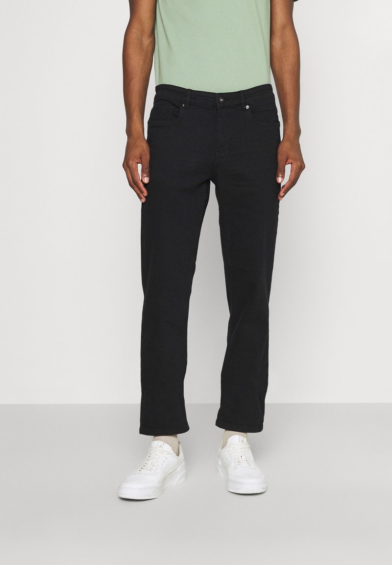 Homme portant un jean droit noir, un t-shirt vert clair et des baskets blanches, debout devant un fond uni.