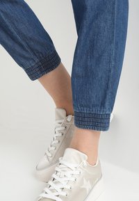 Parties inférieures des jambes portant un pantalon bleu à bords élastiques et des baskets blanches avec un motif étoile sur le côté, sur un fond uni.