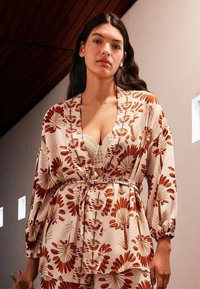 Robe à imprimé floral crème avec des motifs orange et marron. Comprend une taille nouée, des manches longues et une coupe décontractée. Tissu texturé.