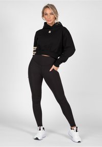 Sudadera corta negra con logo, combinada con leggings negros de cintura alta. La modelo lleva zapatillas deportivas blancas. Textura suave, diseño casual.