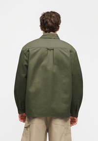 Πουκάμισο ελιάς πράσινο, μακριά μανίκια, μανσέτες με κουμπιά, λεπτομέρεια πίσω πάσου, ίσιο κόψιμο και ύφασμα με ομαλή υφή. Συνδυασμένο με ανοιχτό μπεζ παντελόνι.