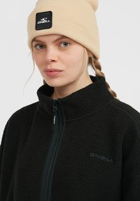 Black fleece jacket with a high collar and front zipper, paired with a beige knitted beanie featuring a black label with the O'Neill logo.