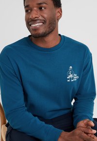 Smiling man wearing a teal long-sleeve shirt with a small white graphic of a cartoon bird holding an ice cream cone on the chest.