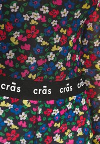 Nahaufnahme eines bunten, floral gemusterten Stoffs mit einem schwarzen elastischen Bund, der "crās" in weißen Buchstaben wiederholt.