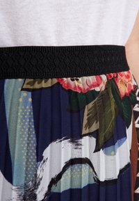 Záhybová sukně s černým tkaným pasem, zdobená květinovým vzorem ve výrazných barvách na tmavém pozadí a jemnými texturovanými vzory.