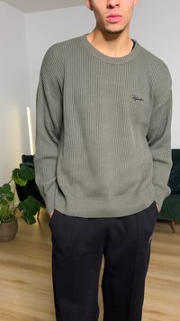 Grønn ribbestrikket genser med rund hals og brodert logo. Kombinert med løse svarte bukser, hender i lommer. Gulvet er av tre.