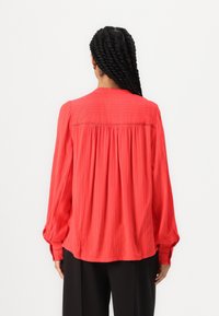 Blusa rossa a maniche lunghe con tessuto strutturato, dettaglio plissettato sul retro e polsini arricciati. Vestibilità ampia, scollatura rotonda e dettagli di cucitura sottili.