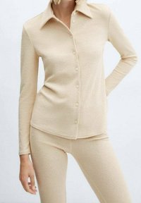 Haut beige à manches longues avec boutons et col, en tissu texturé, assorti à un pantalon ajusté. Les deux articles ont une finition douce.