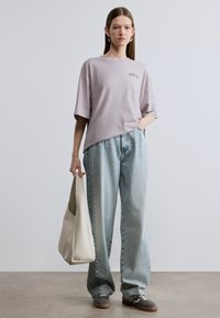 Light purple short-sleeve t-shirt with "PIROUETTES" text. Loose-fitting light blue jeans. Model holds a cream tote bag; grey sneakers featured.