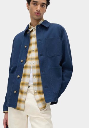 Jonge man die een blauw jasje met knopen draagt over een geel geruit overhemd en witte broek, staand tegen een effen lichte achtergrond.