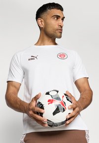 Bijela sportska majica s FC St. Pauli logom i natpisom "KING" u smeškastoj boji. Model drži crno-bijelu nogometnu loptu s crvenim detaljima.