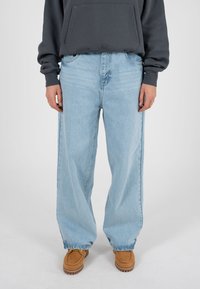 Helesinised laiade säärtega teksad kõrge vöökohaga, millel on tuhmunud pesu ja peen tekstuur, kombineeritud pruunide slip-on kingade ja tumehall kapuutsiga.