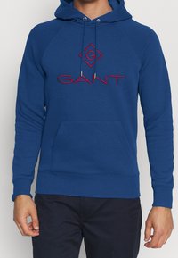 Sudadera azul de algodón, con bolsillo tipo canguro, puños y dobladillo acanalados, con logo bordado en rojo en el pecho y terminales de cordón plateados.