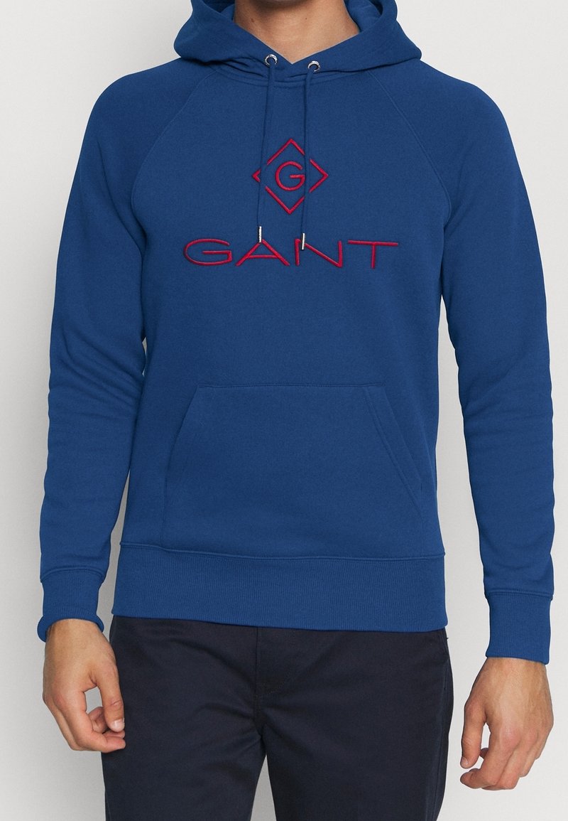 Sudadera azul de algodón, con bolsillo tipo canguro, puños y dobladillo acanalados, con logo bordado en rojo en el pecho y terminales de cordón plateados.