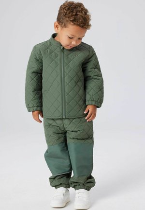 Fiatal gyerek zöld, bélelt dzsekit és hozzáillő, megerősített térdű nadrágot visel, lefelé néz, sima háttér előtt áll.