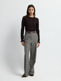 Maglia a maniche lunghe marrone scuro con colletto arricciato e dettagli con bottoni, abbinata a pantaloni a quadri grigi e scarpe in camoscio marroni.