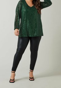 Femme portant un haut à manches longues en sequins vert foncé, des leggings en cuir noir et des sandales à talons hauts noires à lanières, debout.