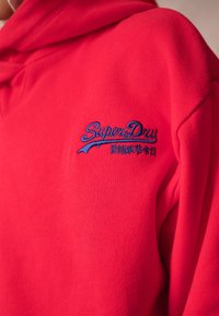 Červená fleecová mikina s kapucí, na hrudi s modrou vyšitou nášivkou "SuperDry". Hladká textura s kapucí a žebrovanými okraji.