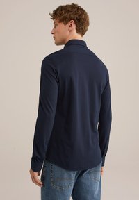 Tamsiai mėlynas ilgomis rankovėmis marškinėliai su klasikiniais apykaklėmis, lygiu medvilniniu audiniu, lieknu siluetu ir susegamais rankogaliais, derinami su šviesiai mėlynais džinsais.