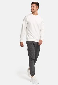 Sudadera blanca de cuello redondo con mangas largas, combinada con pantalones cargo grises que tienen múltiples bolsillos y puños de tobillo con cremallera. Zapatillas blancas.