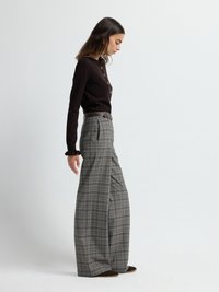 Maglia marrone a maniche lunghe con bottoni e dettaglio a balze, abbinata a pantaloni grigi a quadri con gamba larga. Accentuata da una cintura e scarpe in camoscio.