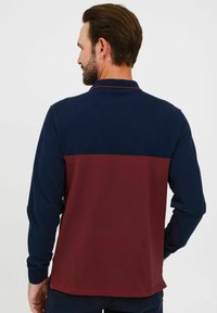 Polo majica dugih rukava s gornjom polovinom u mornarsko plavoj boji i donjom polovinom u tamnocrvenoj boji. Ima ovratnik i bočne proreze na rubu s rebrastim manžetama.