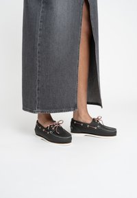 Scarpe da barca nere con suole bianche, superiore in pelle, dettagli rosa e occhielli in metallo. Indossate con una gonna di jeans grigia con una spaccatura laterale.