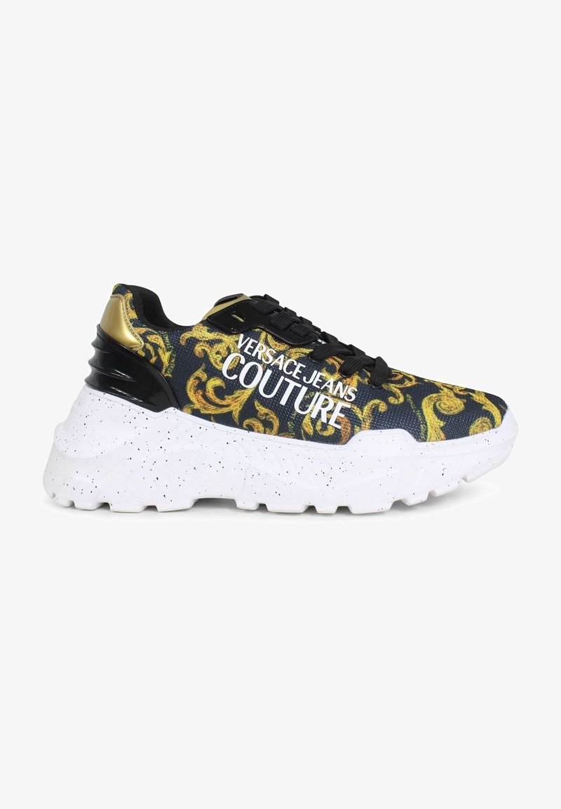 Sneaker Versace Jeans Couture avec un dessus noir orné d'un motif baroque doré, une semelle blanche tachetée et un logo prononcé sur le côté.