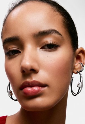 Primo piano di una donna con pelle liscia che indossa grandi orecchini a cerchio argentati con piccoli ciondoli a forma di fiore.