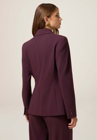 Donna con lunghi capelli castani, che indossa un abito sartoriale viola scuro e grandi orecchini d'oro ornamentali, rivolta di lato su uno sfondo semplice.