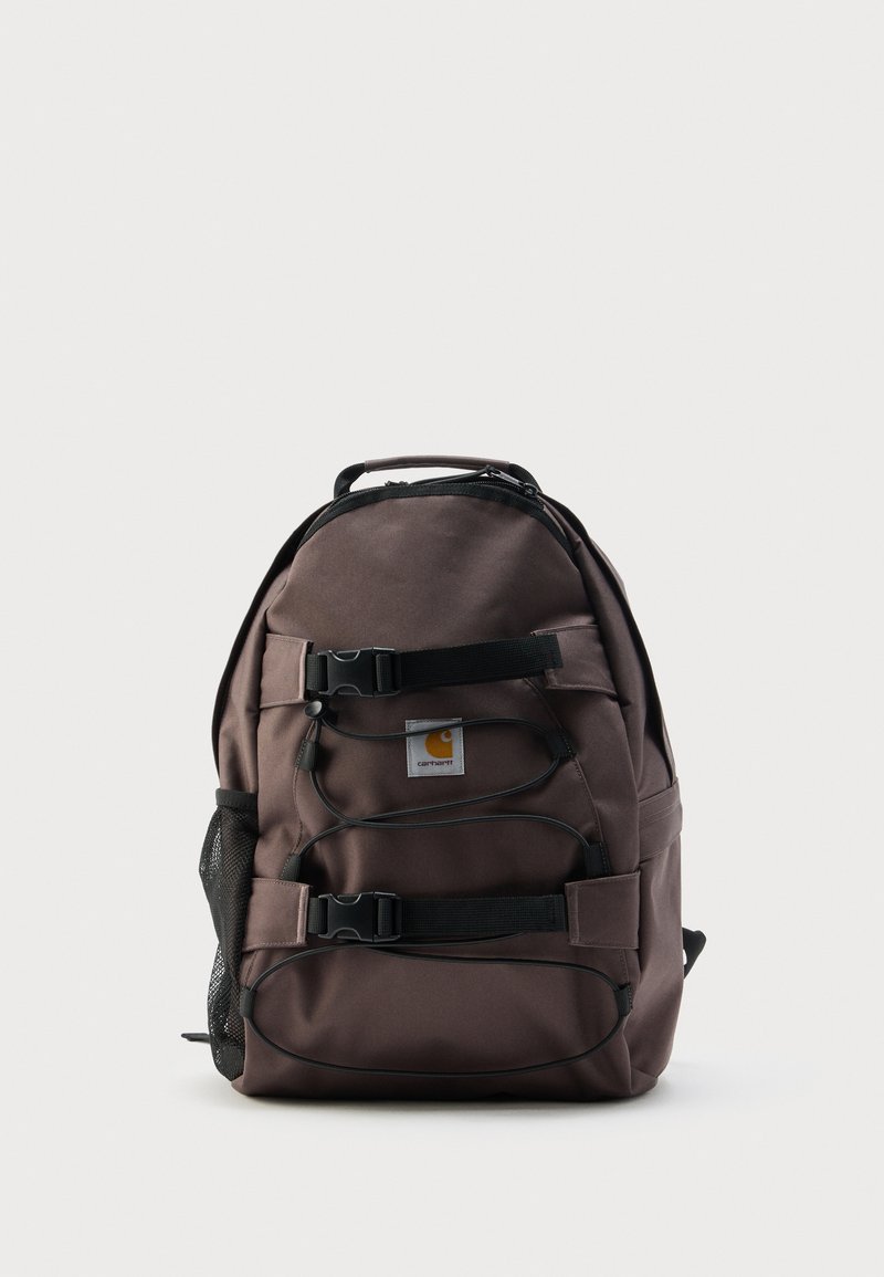 Bruine Carhartt-rugzak met zwarte riemen, gespen, elastische koorden en zijvak van mesh, op een effen witte achtergrond.